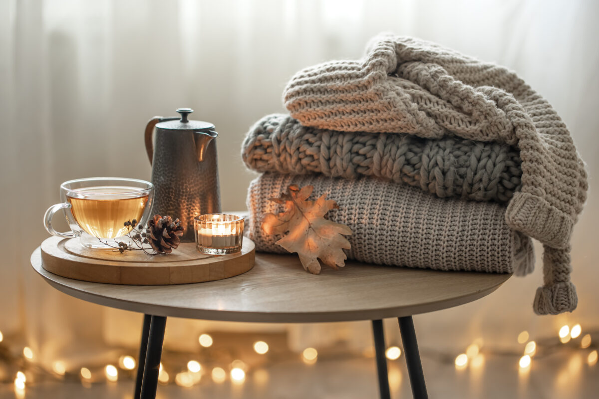 Artigos para decoração no inverno, dispostos em cima de uma mesa. Uma chávena e bule de chá junto a uma vela e mantas de tecidos quente